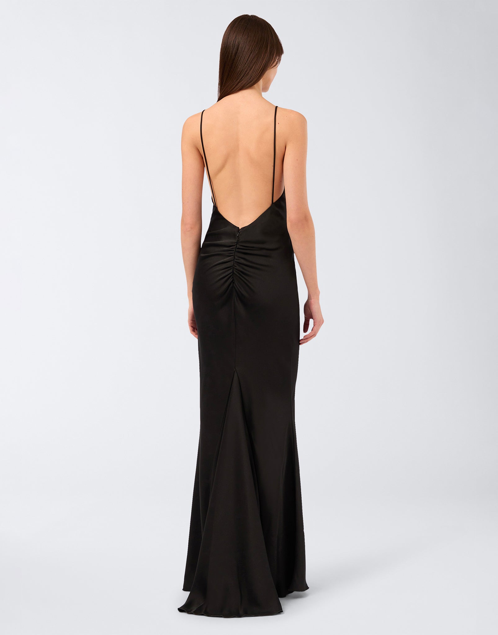 Rear view: Woman wearing Ninfea Maxi Dress in black crepe satin, showcasing a low open back with delicate straps, draped detail, and flowing maxi skirt.