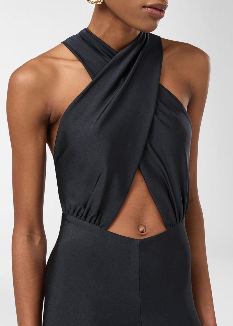 Frontal view of the Hola Jumpsuit showing a black shiny lycra halterneck with overlapping straps, a triangular front cut-out, and slim, body-skimming legs.