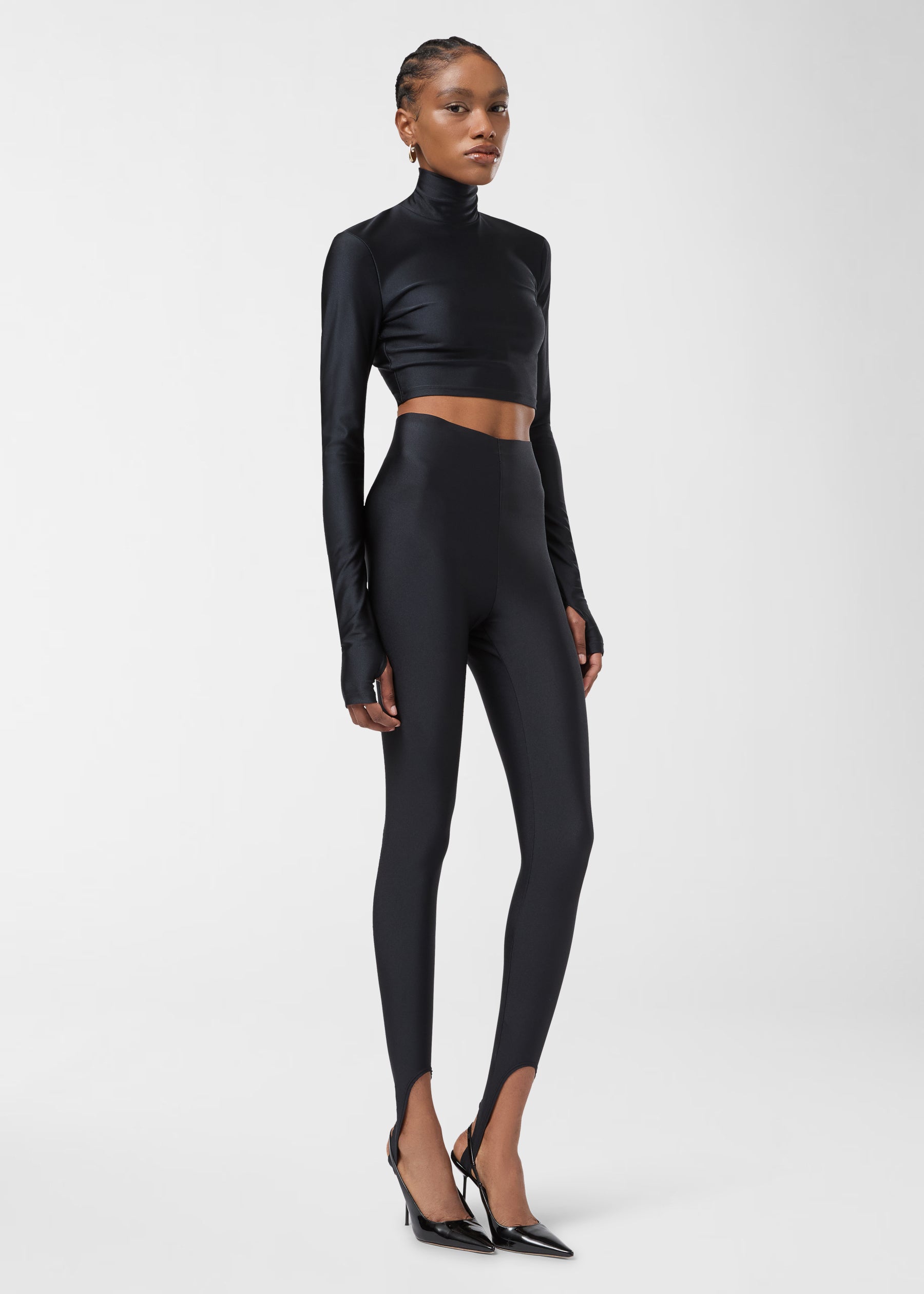 Frontal view of New Holly Stirup Leggings showing black shiny full-length stirrup leggings with a very high raw hem waist, tight fit, and foot straps on a model.