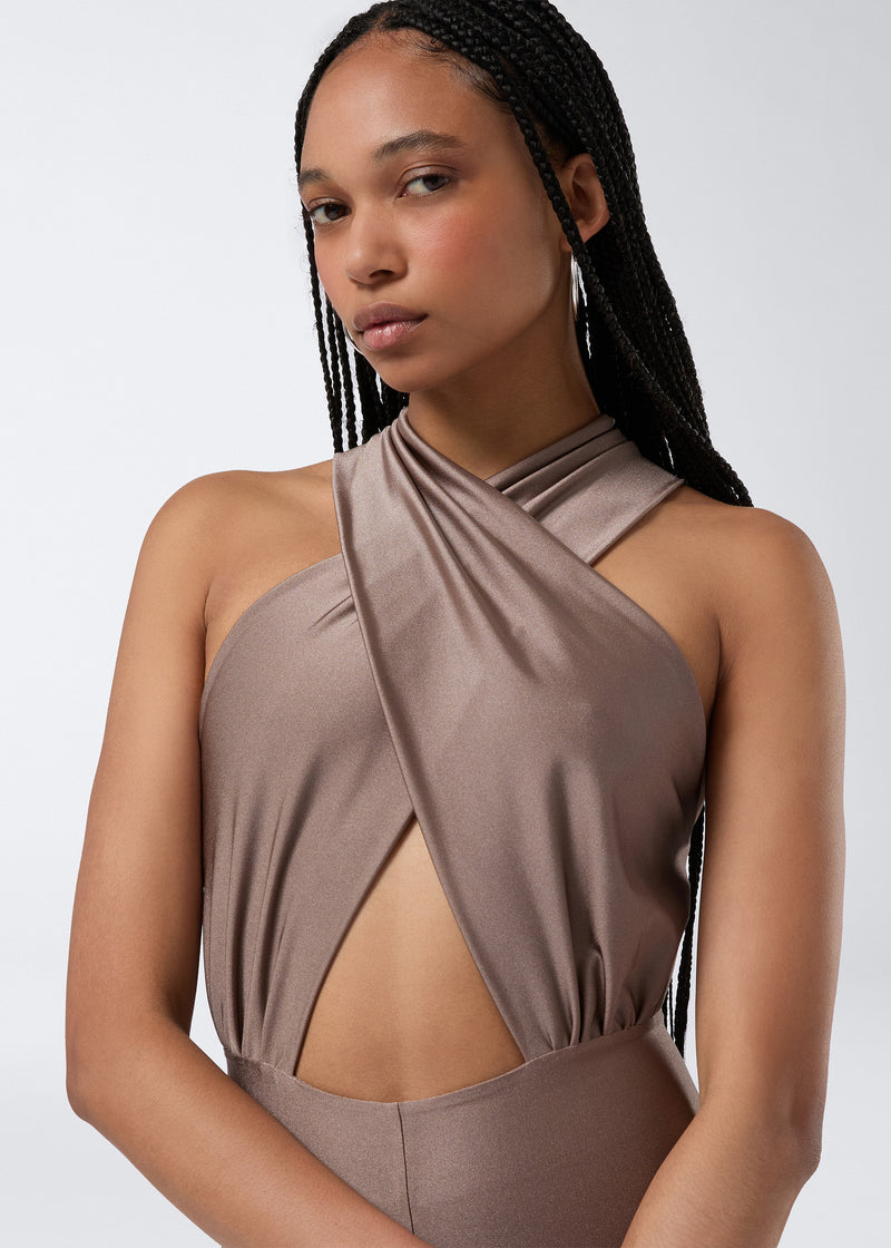 
                          Frontal view of a model wearing the Hola Jumpsuit in greige shiny lycra, showcasing the crossover halterneck with a triangular front cut-out detail.
                        