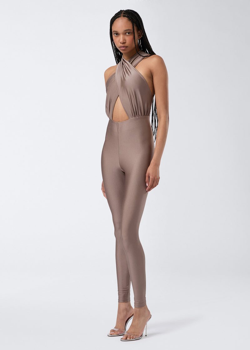 
                          Frontal view of a model wearing the Hola Jumpsuit in greige shiny lycra, featuring a crossover halterneck, triangular cut-out detail, and skin-tight full-length leggings.
                        