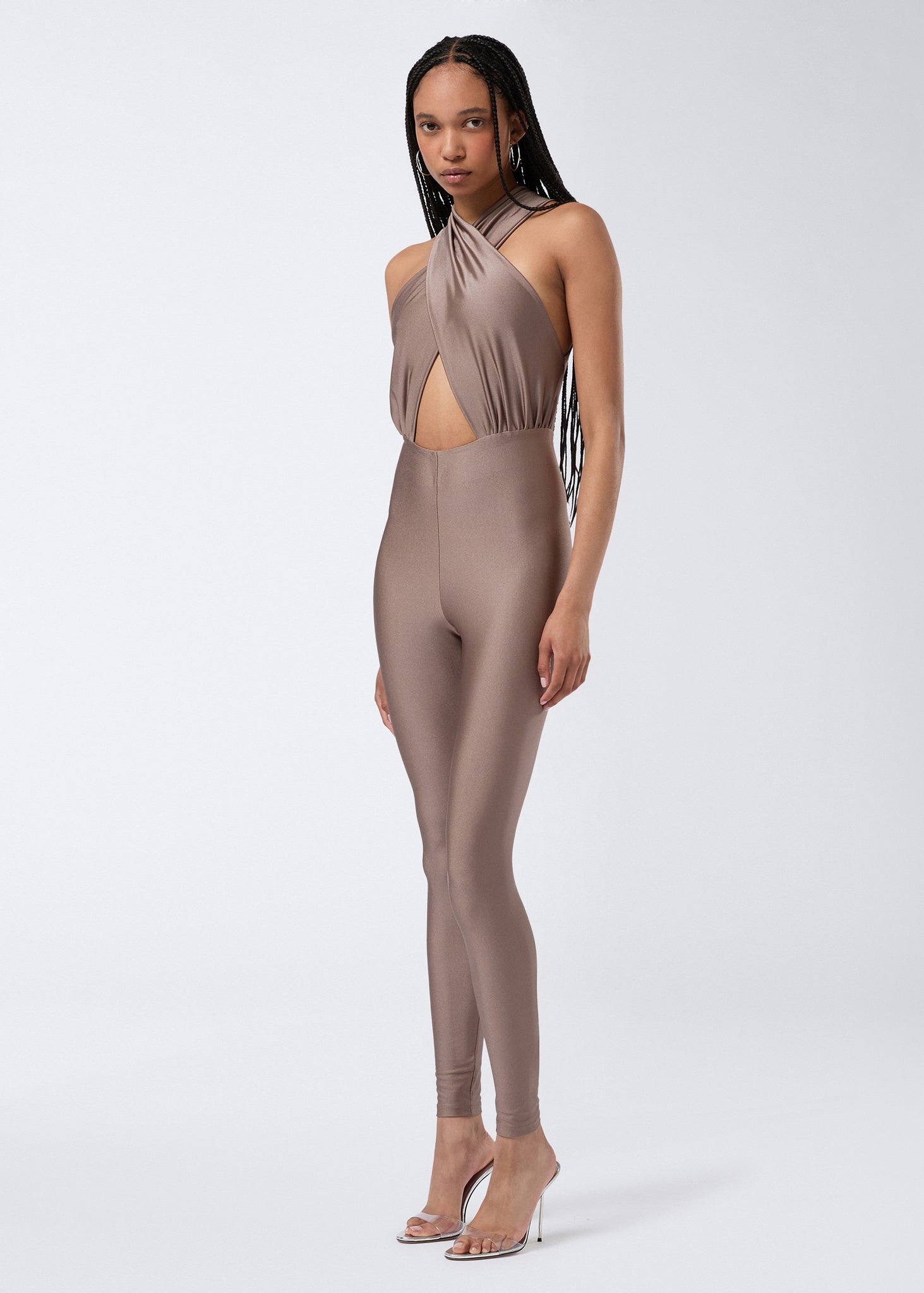 Frontal view of a model wearing the Hola Jumpsuit in greige shiny lycra, featuring a crossover halterneck, triangular cut-out detail, and skin-tight full-length leggings.