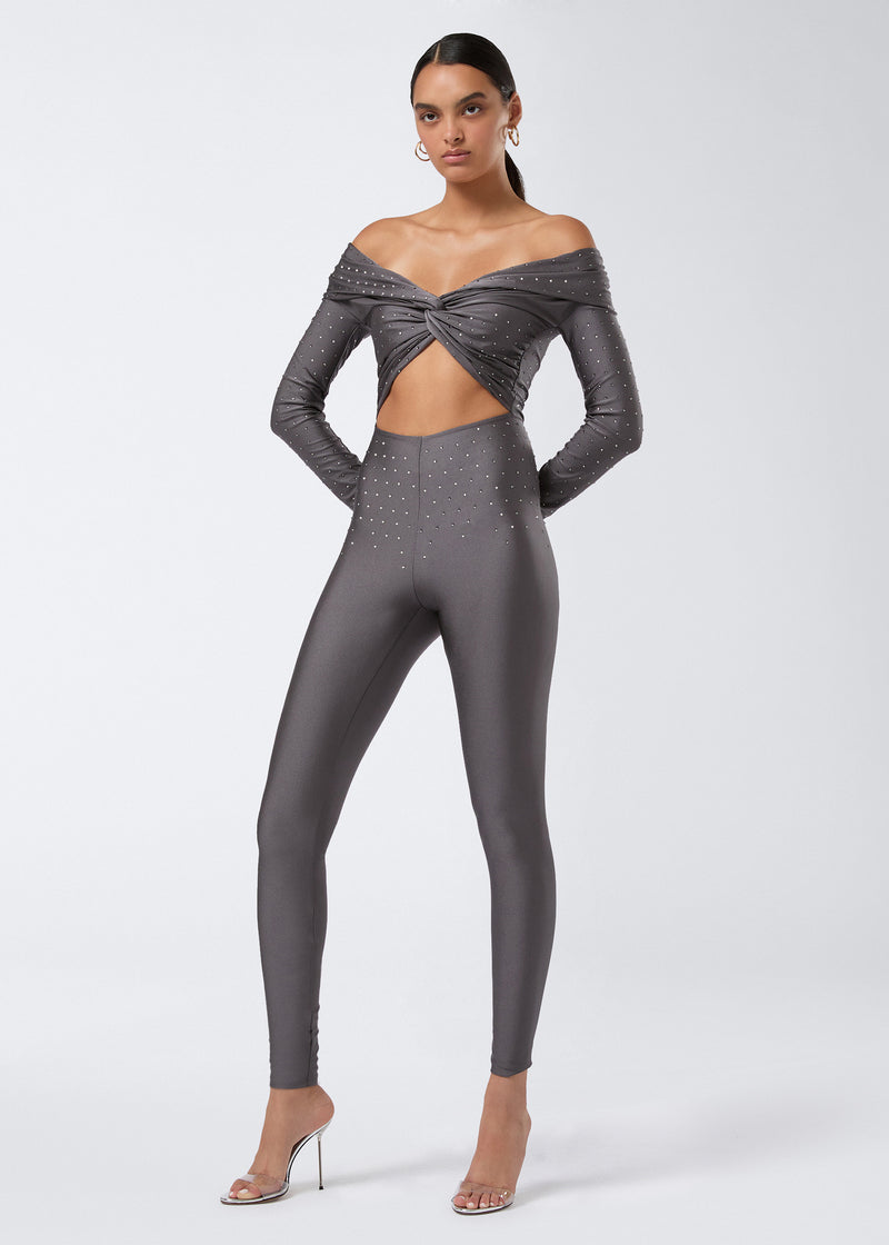 
                          Frontal view of the Kendall Crystal Jumpsuit in ardesia shiny lycra with off-the-shoulder neckline, chest cut-out, knot detail, full-length leggings, and crystal embellishments.
                        