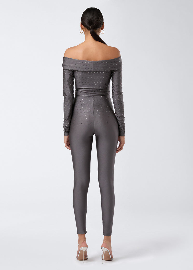 
                          Rear view of a woman wearing the Kendall Crystal Jumpsuit in shiny ardesia lycra, adorned with crystals, off-the-shoulder neckline and full-length leggings.
                        