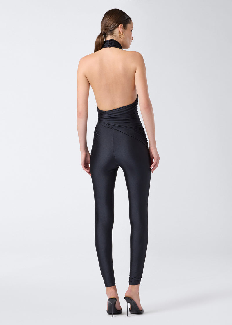 
                          Rear view: Model wears the Tatiana Jumpsuit in black shiny lycra, showcasing a sleeveless halterneck design, deep open back, and full-length leggings.
                        