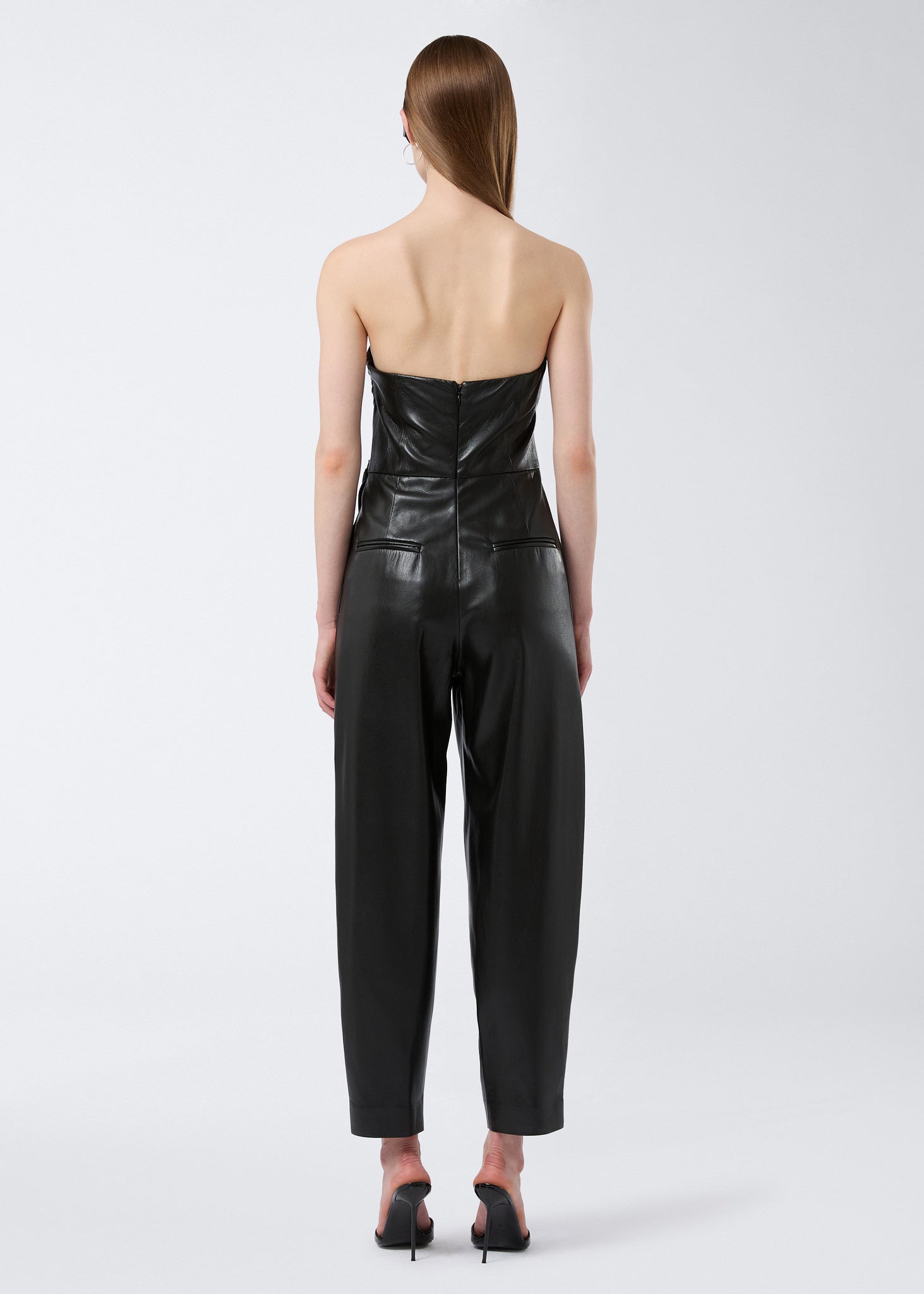 Rear view of a model wearing the Yana Jumpsuit in black vegan leather, showcasing the strapless bustier neckline, cinched waist, and zip fastening at the back.
