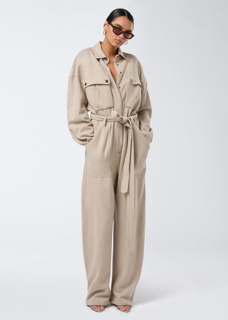 
                          Frontal view of a woman wearing the Xandra Jumpsuit in sand-colored soft felted jersey, featuring a pointed collar, buttoned patch pockets, elasticated waist with belt, and long sleeves.
                        