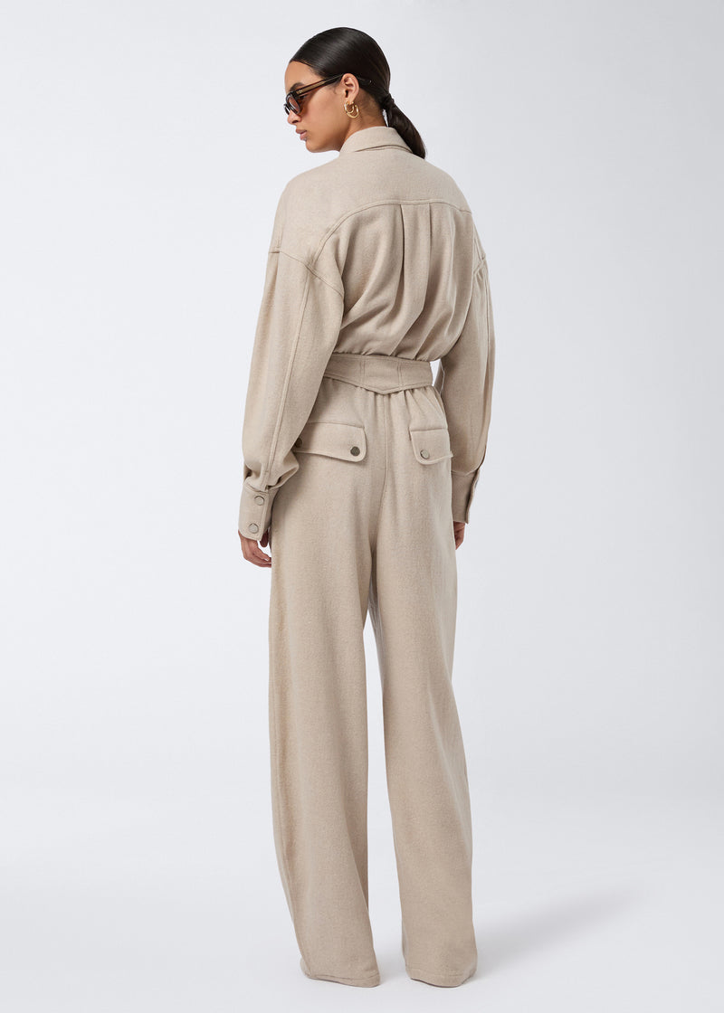 
                          Rear view of a woman wearing the Xandra Jumpsuit, showcasing a sand-colored, felted jersey jumpsuit with a pointed collar, belted waist, and buttoned patch pockets.
                        