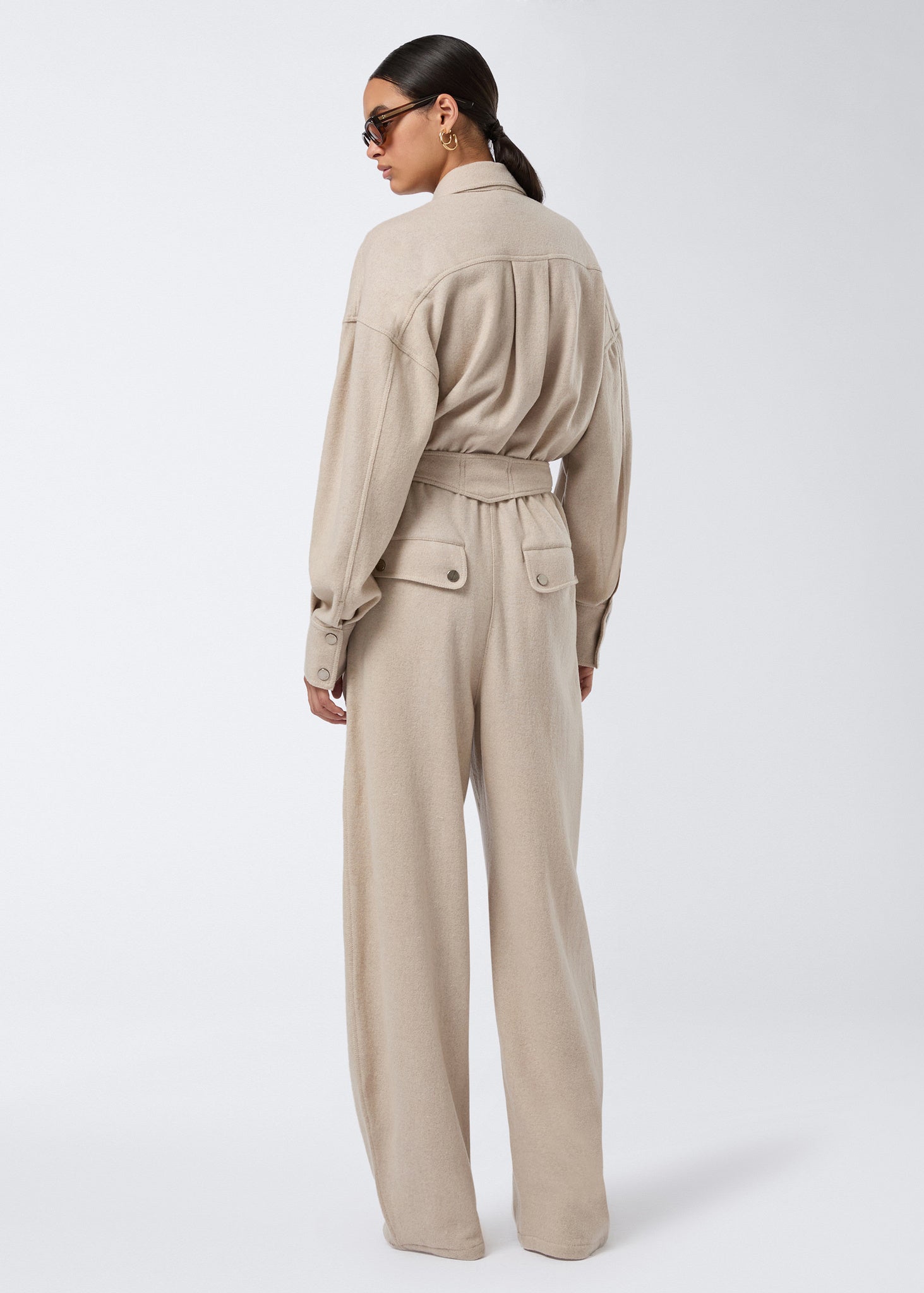 Rear view of a woman wearing the Xandra Jumpsuit, showcasing a sand-colored, felted jersey jumpsuit with a pointed collar, belted waist, and buttoned patch pockets.