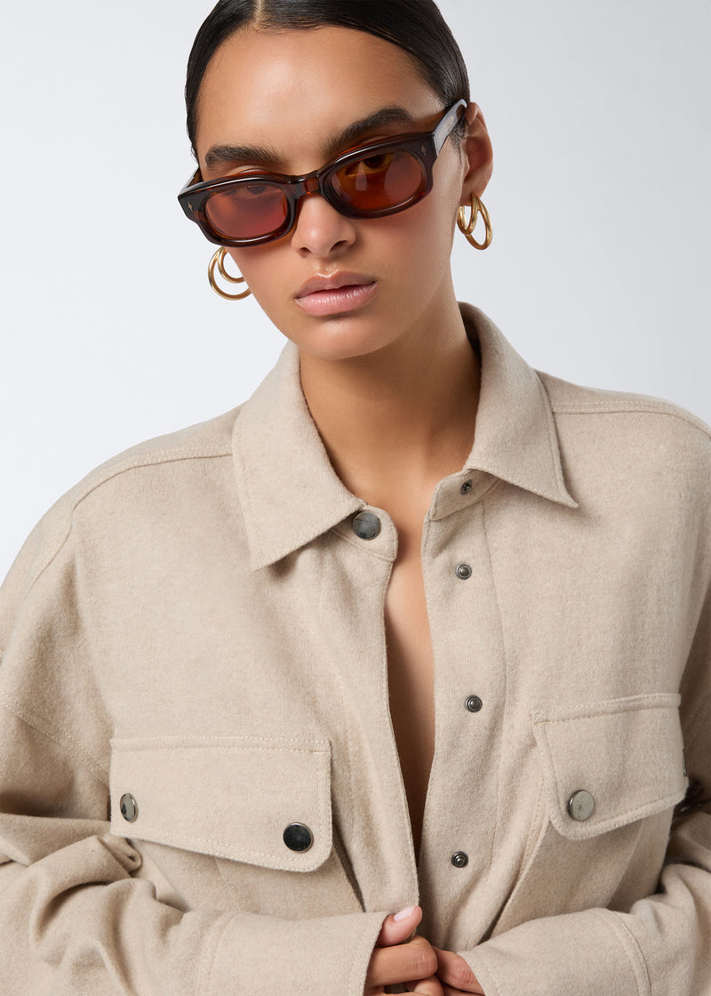 
                          Frontal detail view of the Xandra Jumpsuit in sand-colored soft felted jersey, highlighting the pointed collar, snap buttons, and buttoned patch pockets.
                        