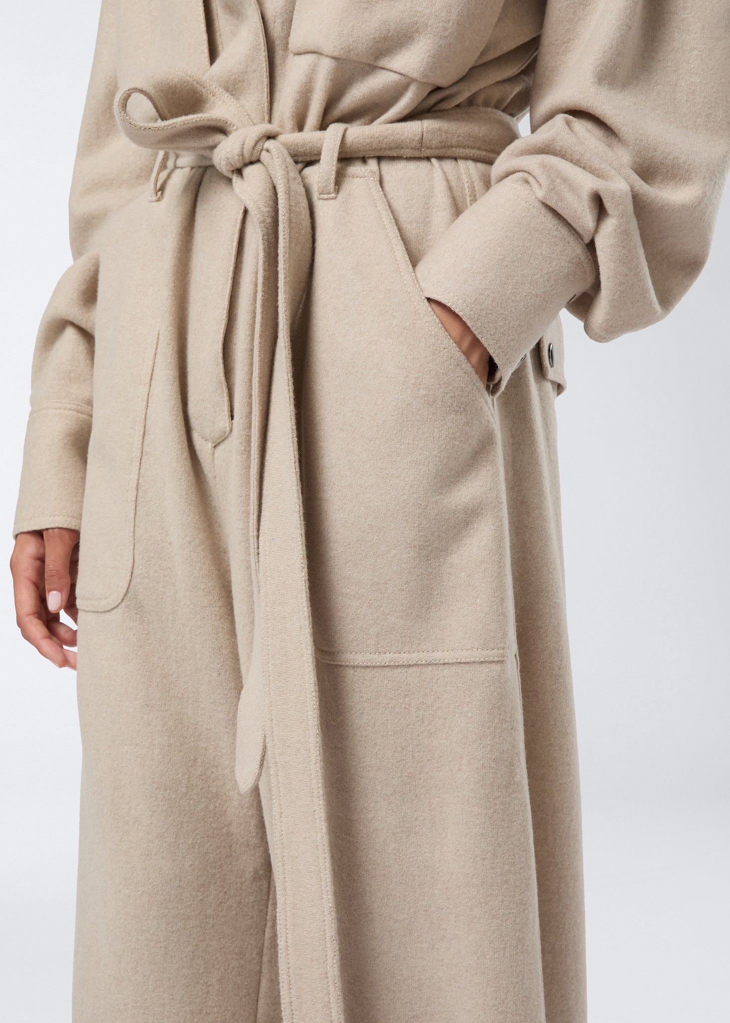 Detail view shows a woman&#39;s hand in the pocket of the sand-colored Xandra Jumpsuit, highlighting its felted jersey fabric, belt, and large patch pockets.