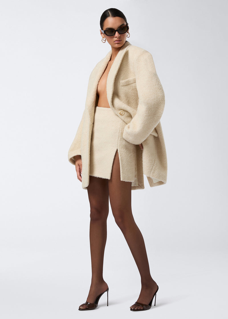 
                          Frontal view of a woman wearing the Gioia Mini Skirt in soft ivory wool twill, high-waisted with a thigh side slit, styled with a matching oversized ivory jacket, sheer black tights, and heels.
                        