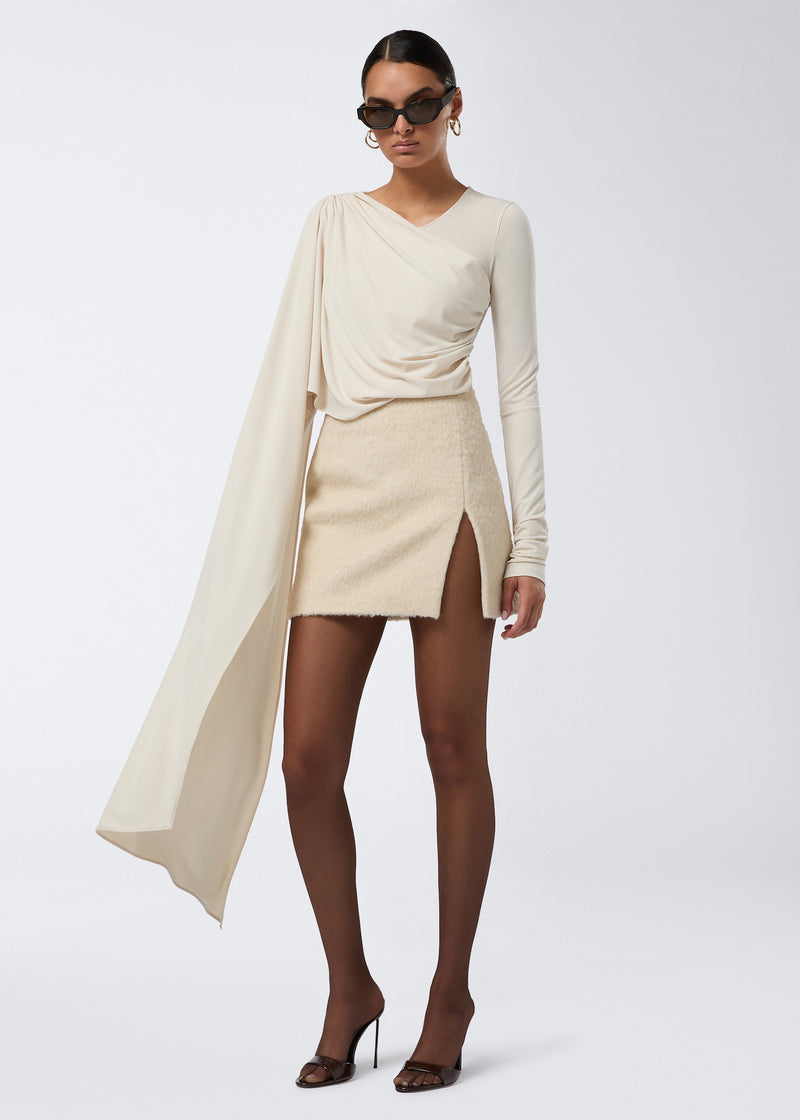 
                          Frontal view of a woman wearing the Gioia Mini Skirt in soft ivory wool twill. The high-waisted skirt features a thigh-high side slit and a secure zip fastening.
                        