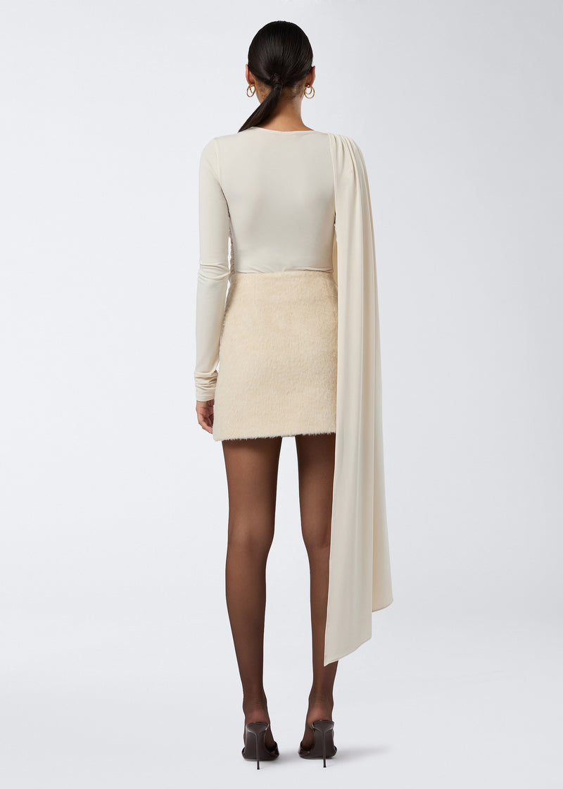 
                          Rear view of a model wearing the Gioia Mini Skirt, a soft ivory wool twill high-waisted mini skirt, paired with sheer black tights and heels.
                        