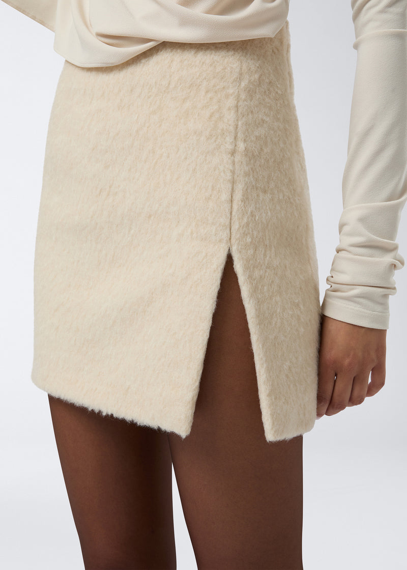 
                          Frontal view of a woman wearing the Gioia Mini Skirt in soft ivory wool twill, featuring a high waist, side thigh slit, and textured fabric finish.
                        
