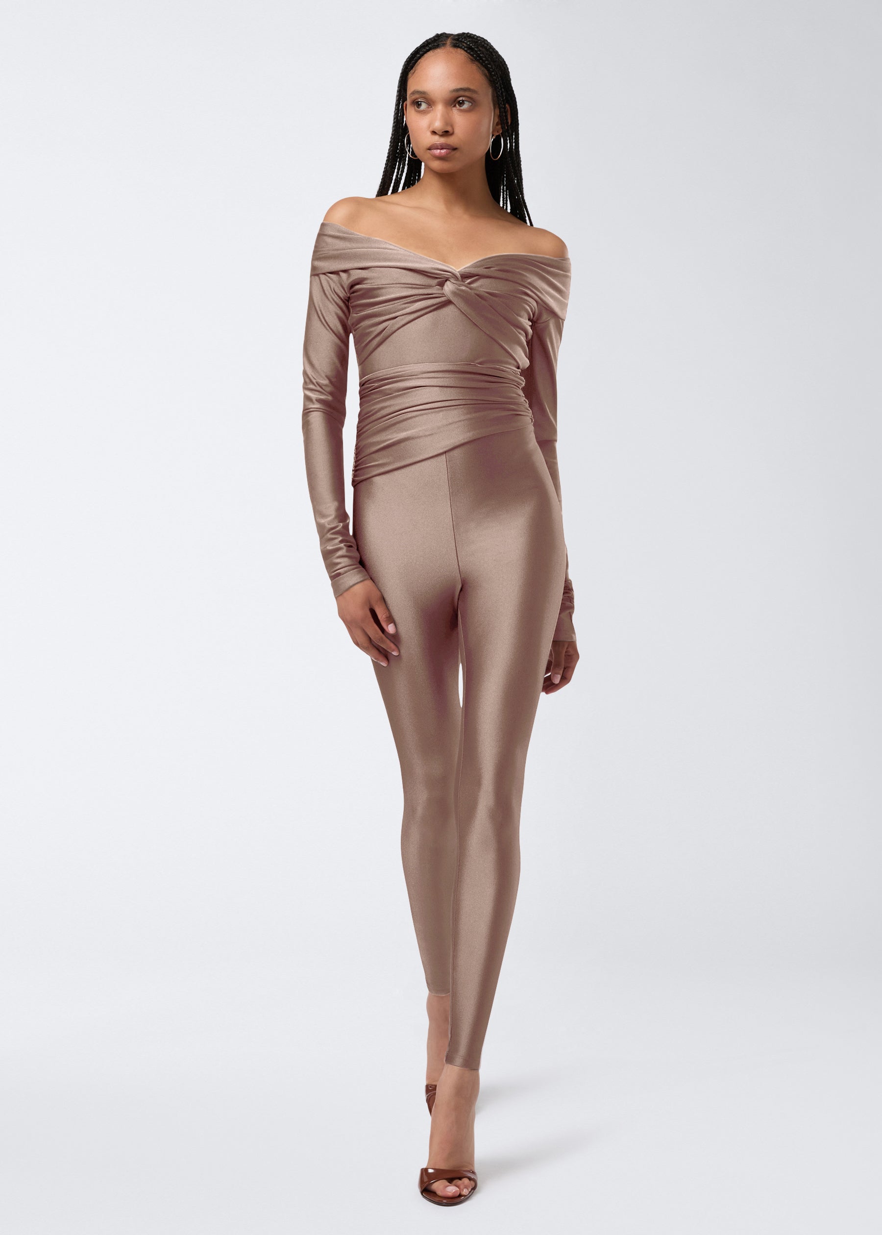 Frontal view of a model wearing Vicky Leggings in a greige shiny lycra fabric, featuring high waist, full-length fit, and draped detail around the waist.
