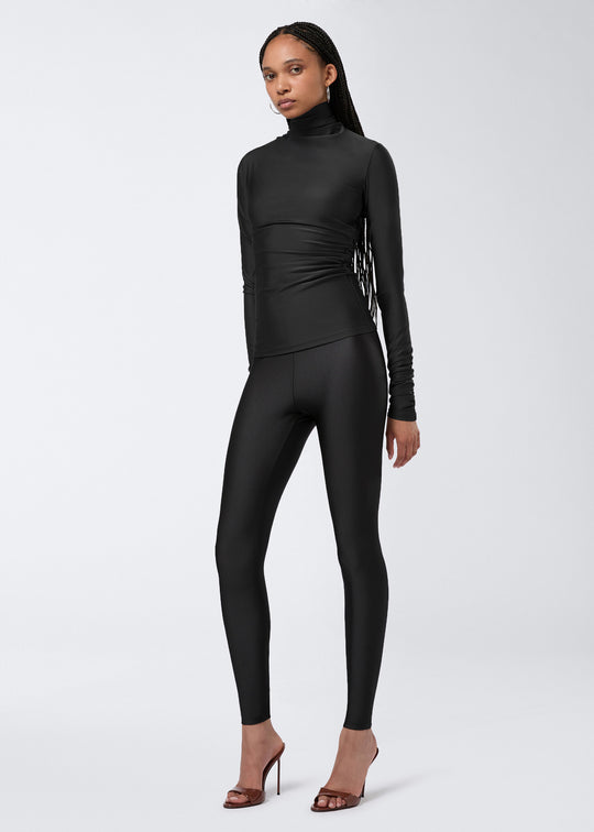 Frontal view of a woman wearing the Oleandra Top, a black shiny lycra turtleneck top with long sleeves and draping at the waist, paired with black leggings.