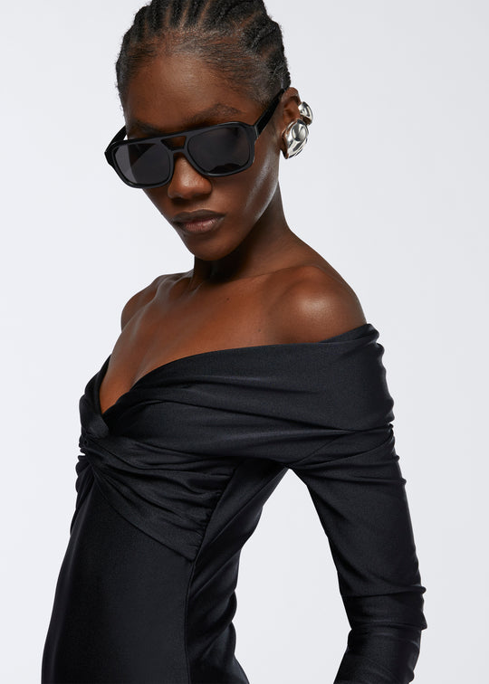 Frontal view of a model wearing the Kendall Bodysuit in black shiny lycra, featuring an off-shoulder v-neckline with a knotted detail and long sleeves.