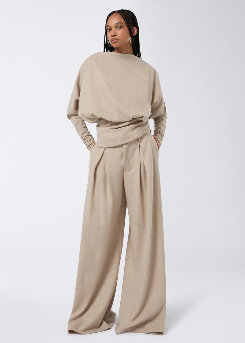 
                          Frontal view of a model wearing the Wisteria Top, a sand-colored felted jersey t-shirt with long sleeves, boat neckline, draped waistband, and a relaxed fit.
                        