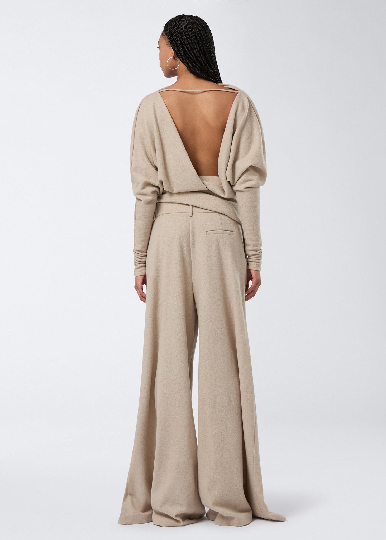 Rear view showing a person wearing the sand-colored Wisteria Top with a plunging open back, long sleeves, draped waistband, and wide-leg pants.