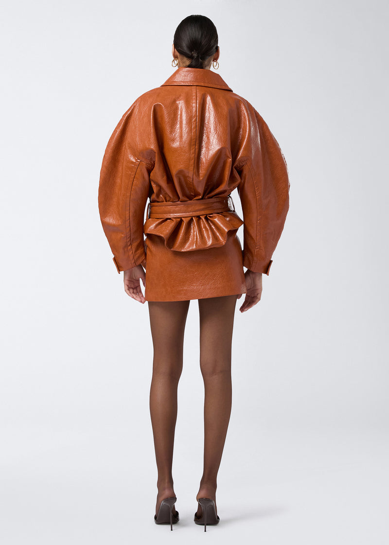 
                          Rear view of a woman wearing the Zarina Coat, a cropped cuoio-shade embossed eco leather coat with large collar, padded structure, and matching belt.
                        