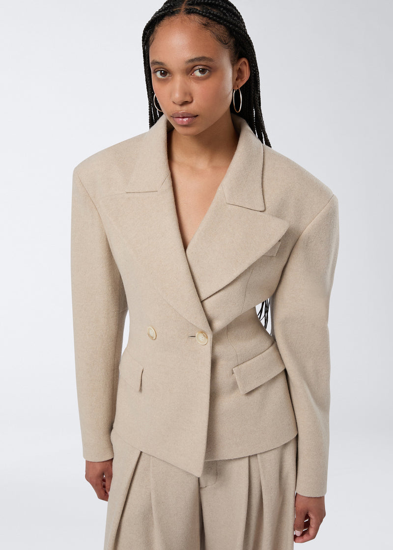 
                          Frontal view of a model wearing the Tillie Blazer in sand-colored felted jersey, featuring a double-breasted closure, wide lapel, and flap pockets.
                        