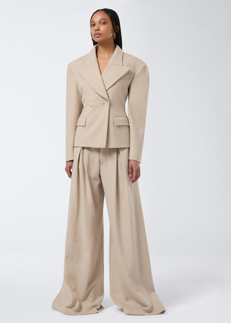 
                          Frontal view of a model wearing the Tillie Blazer in sand-colored felted jersey, featuring a double-breasted closure, wide lapel, padded shoulders, and flap pockets.
                        