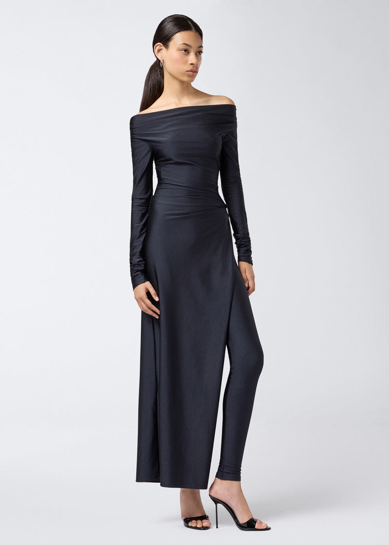 
                          Frontal view of a model wearing the Wallis Jumpsuit, a black shiny lycra off-the-shoulder jumpsuit with long sleeves, draped waist, and high leg slit revealing inner leggings.
                        