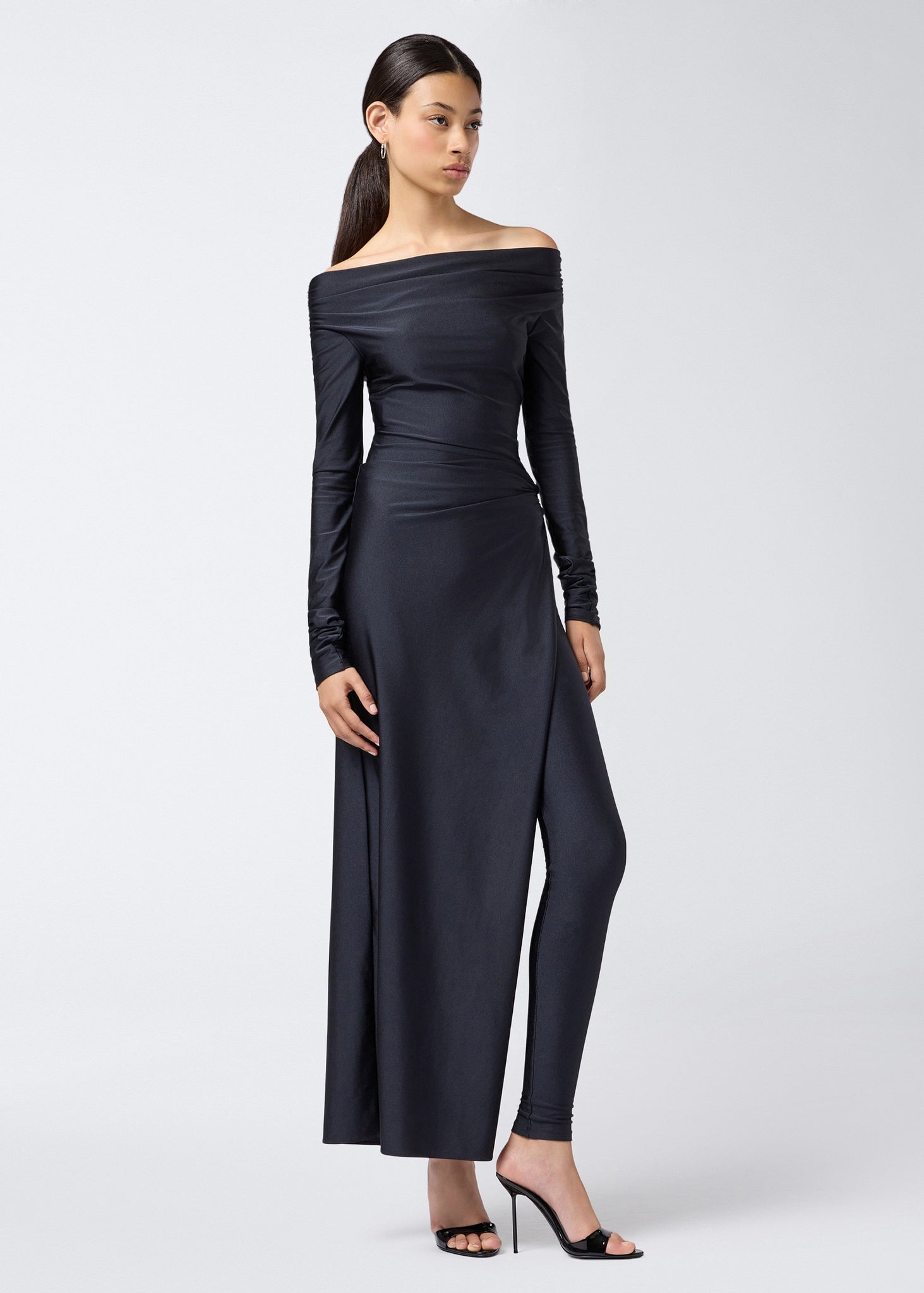 Frontal view of a model wearing the Wallis Jumpsuit, a black shiny lycra off-the-shoulder jumpsuit with long sleeves, draped waist, and high leg slit revealing inner leggings.