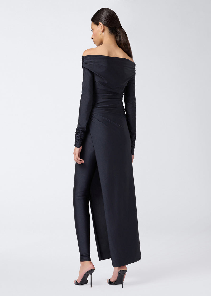 
                          Rear view of a woman wearing the Wallis Jumpsuit in shiny black lycra, featuring off-the-shoulder long sleeves, draped waist, and a high leg slit revealing inner leggings.
                        