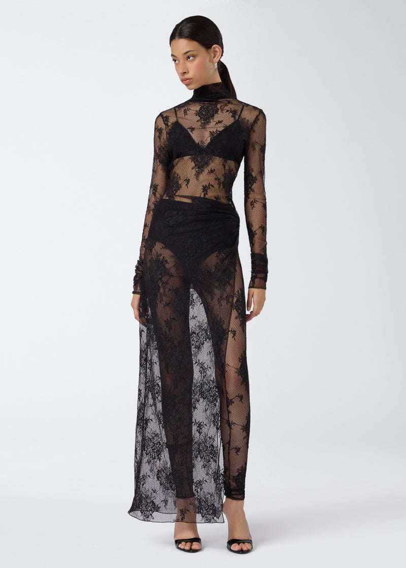 
                          Frontal view shows a model wearing the Tessa Jumpsuit, a black lace, sheer, long-sleeve women’s jumpsuit with turtleneck, knot-draped waist, and slit maxi skirt.
                        