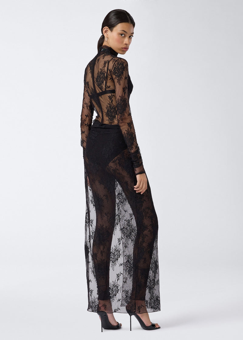 
                          Rear view of a woman wearing the Tessa Jumpsuit, featuring sheer black lace, long sleeves, open back, and full-length leggings beneath a maxi skirt with a high slit.
                        