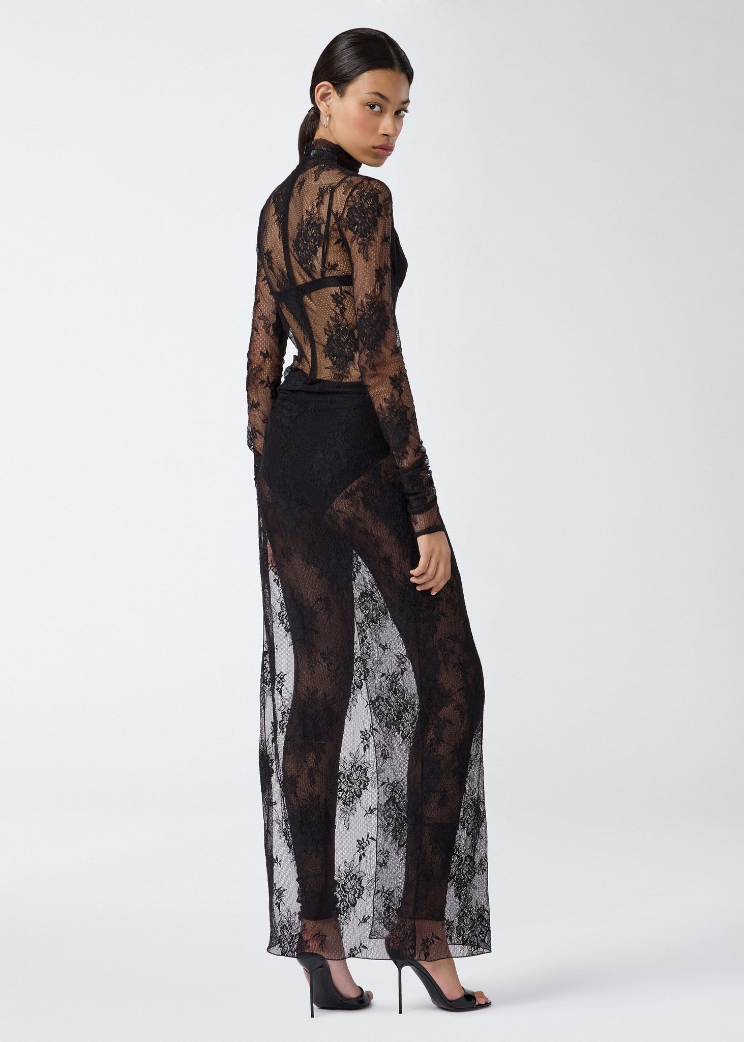 Rear view of a woman wearing the Tessa Jumpsuit, featuring sheer black lace, long sleeves, open back, and full-length leggings beneath a maxi skirt with a high slit.