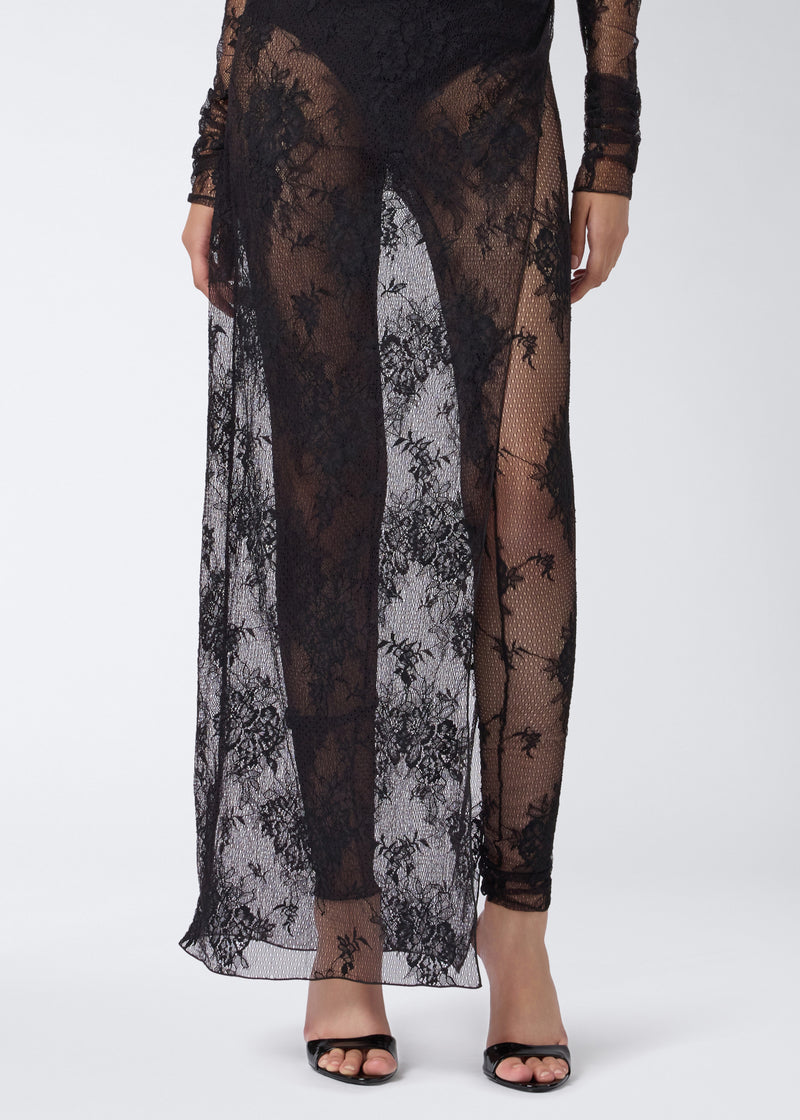 
                          Detail view of the Tessa Jumpsuit showing sheer black lace fabric with floral patterns, a deep skirt slit, and long sleeves over full-length leggings.
                        