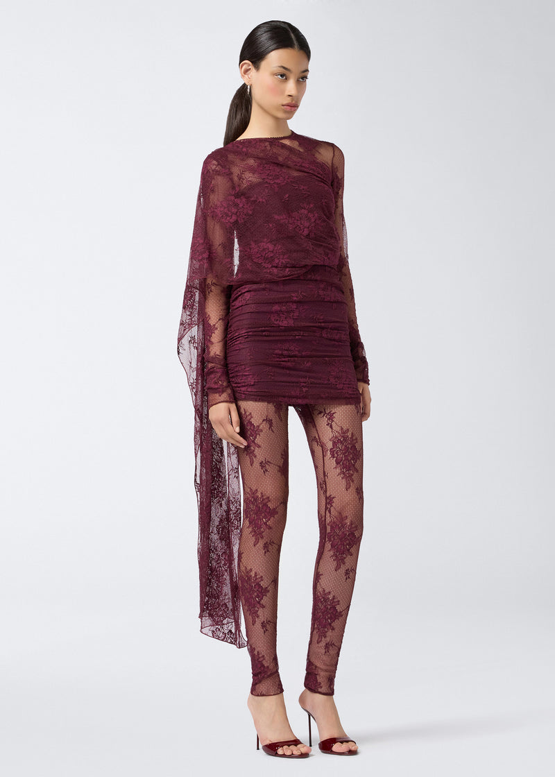 
                          Frontal view of a woman wearing the Xenia Mini Dress in cabernet lace, featuring sheer long sleeves, a cape detail over the shoulder and back, built-in straps, ruching, and a short, fitted silhouette.
                        
