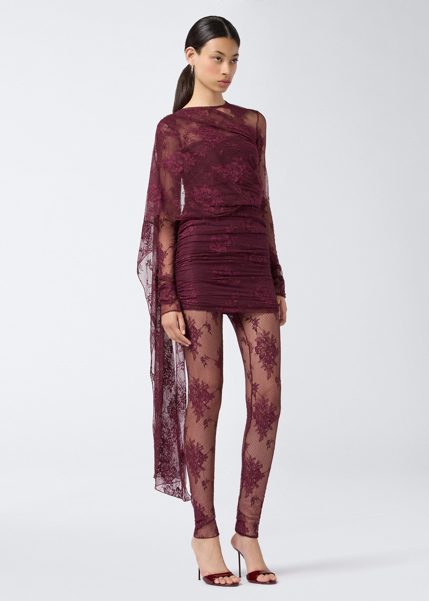 Frontal view of a woman wearing the Xenia Mini Dress in cabernet lace, featuring sheer long sleeves, a cape detail over the shoulder and back, built-in straps, ruching, and a short, fitted silhouette.