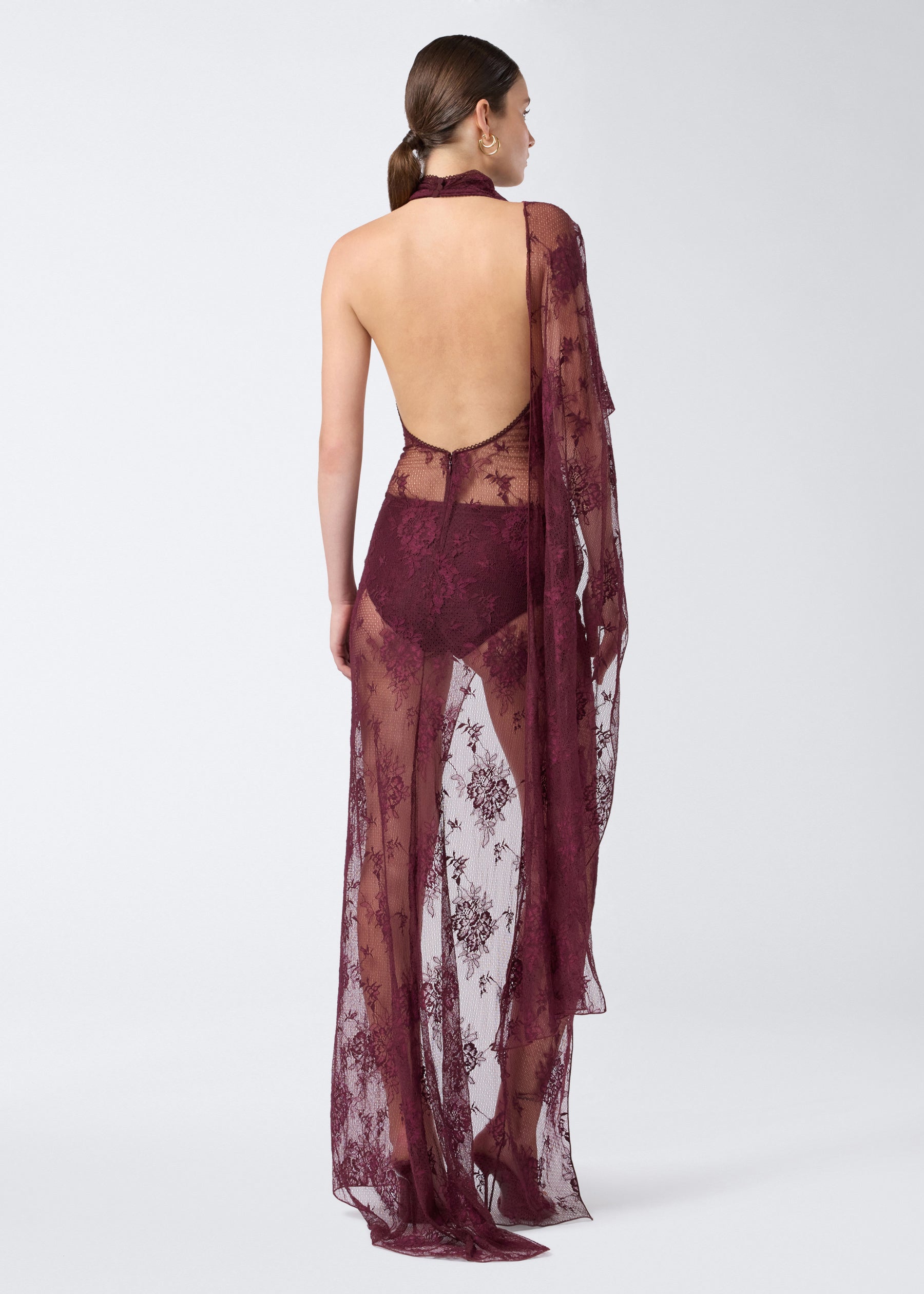 Rear view shows a model wearing the Waverly Maxi Dress in cabernet lace, featuring a sheer finish, backless design, draped cape over one shoulder, and floor-length skirt.