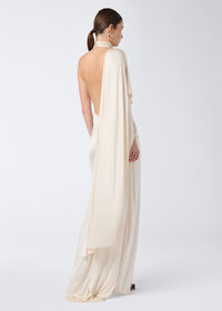 Rear view: Model wears Waverly Maxi Dress in ivory shiny jersey with a dramatic cape effect draping over one shoulder and an open back.