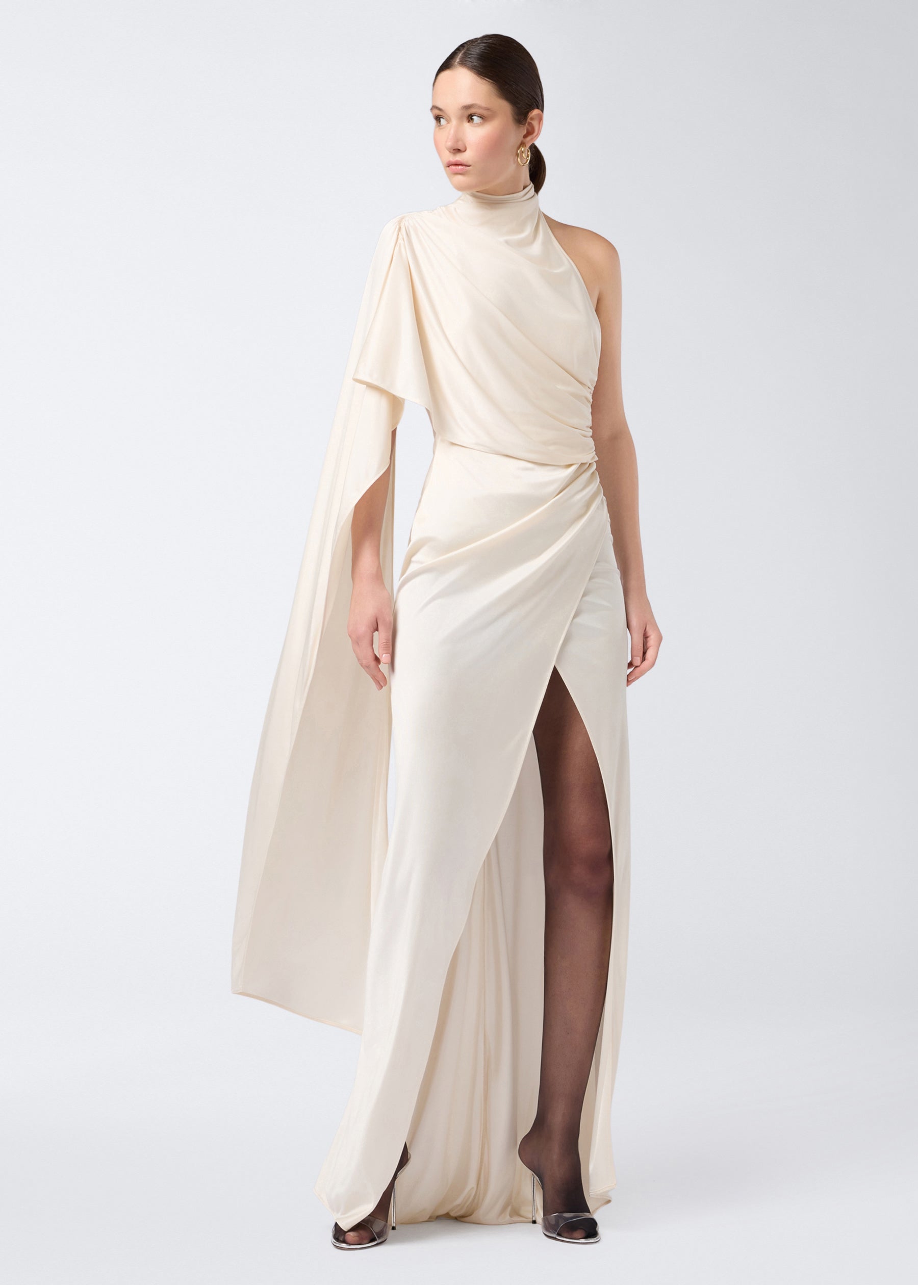 Frontal view of the Waverly Maxi Dress in ivory shiny jersey, featuring a long shoulder cape, turtleneck, floor-length skirt, and thigh-high slit.