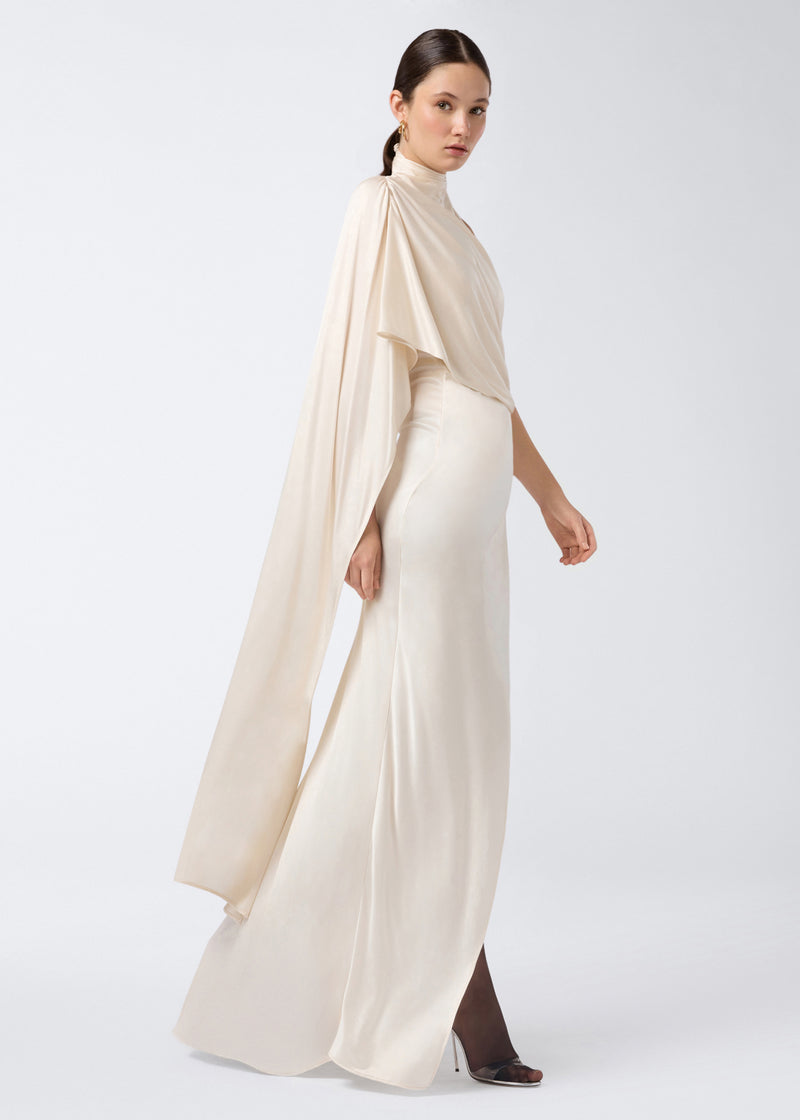 
                          Side view showing a model wearing the Waverly Maxi Dress in ivory shiny jersey, featuring a long cape draped over the shoulder and back, elongated silhouette, and floor-length slit skirt.
                        