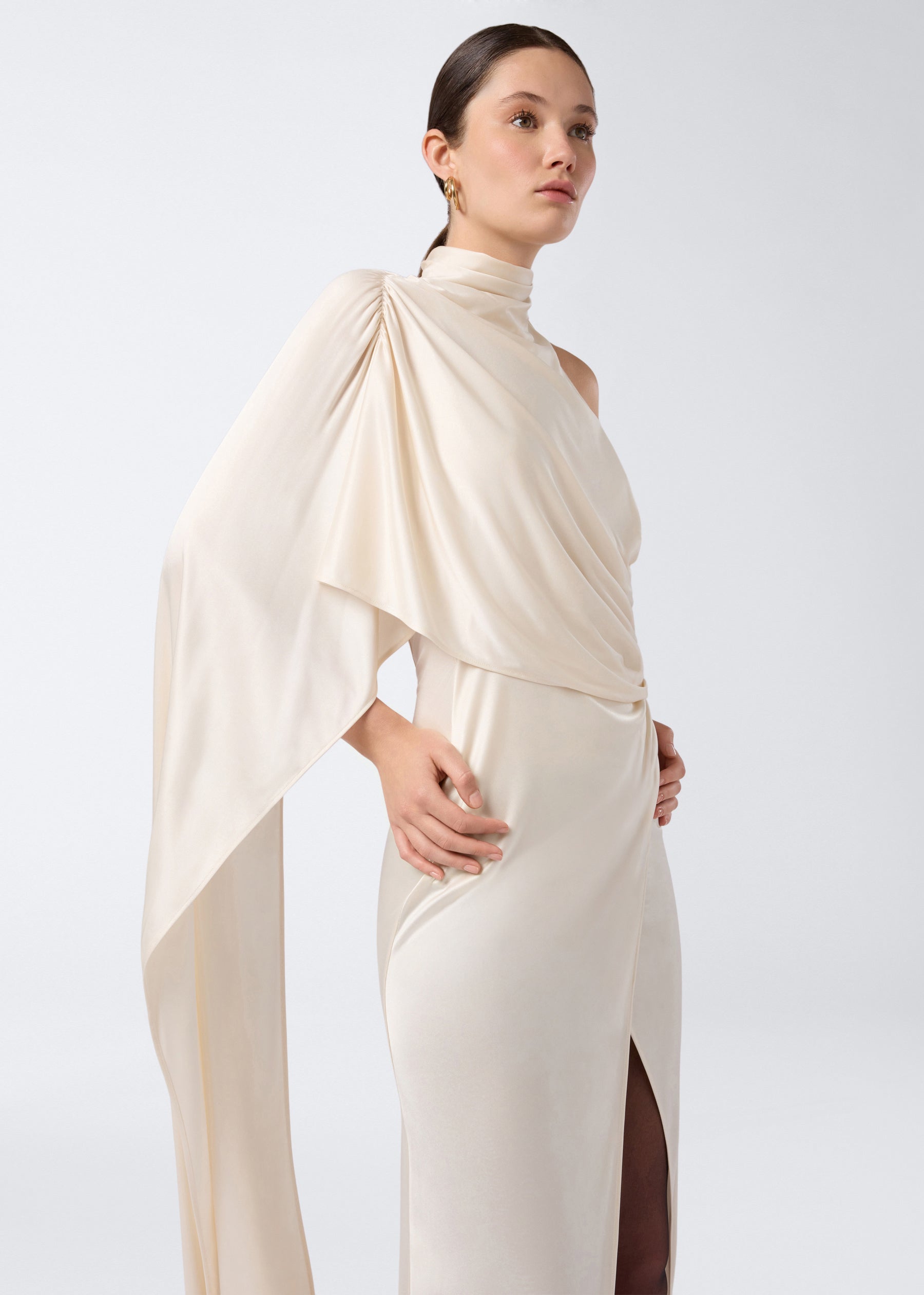 Frontal view shows a woman wearing the Waverly Maxi Dress in ivory shiny jersey, featuring a single-shoulder cape draping down her arm and a high slit.