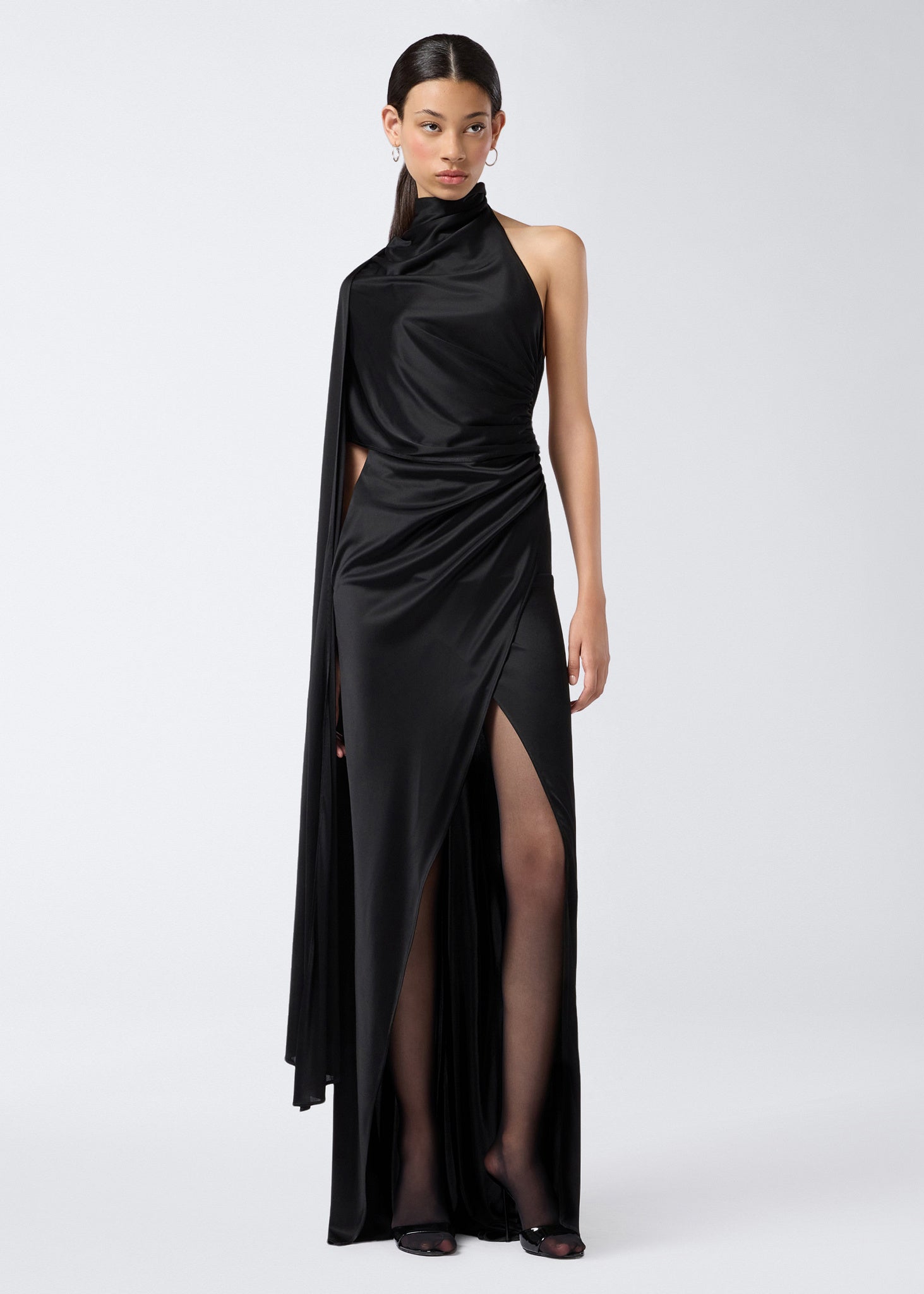 Frontal view of the Waverly Maxi Dress, a black shiny jersey women’s dress with a dramatic cape extending over one shoulder, turtleneck, and high slit skirt.
