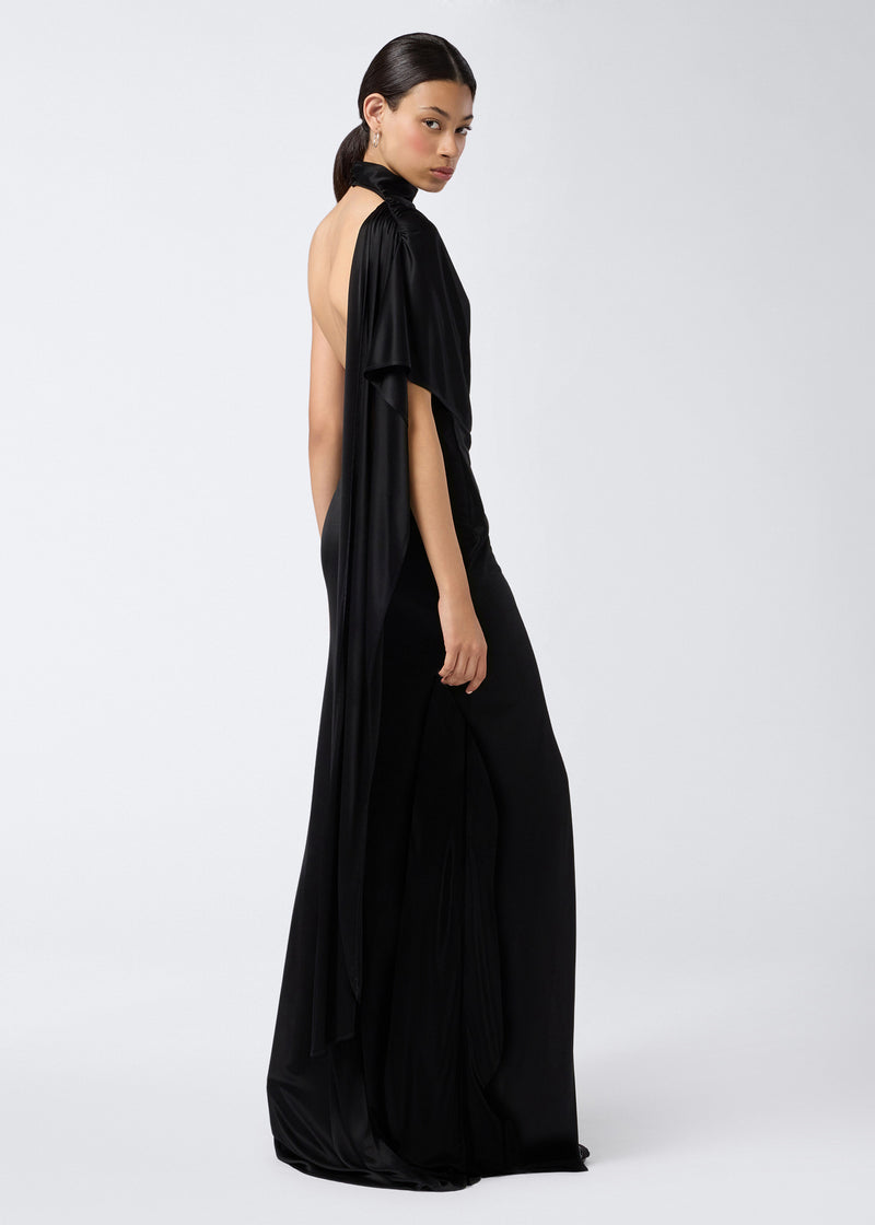 
                          Side view of a woman wearing the Waverly Maxi Dress in black shiny jersey, featuring a dramatic cape effect over one shoulder, turtleneck, and high slit.
                        