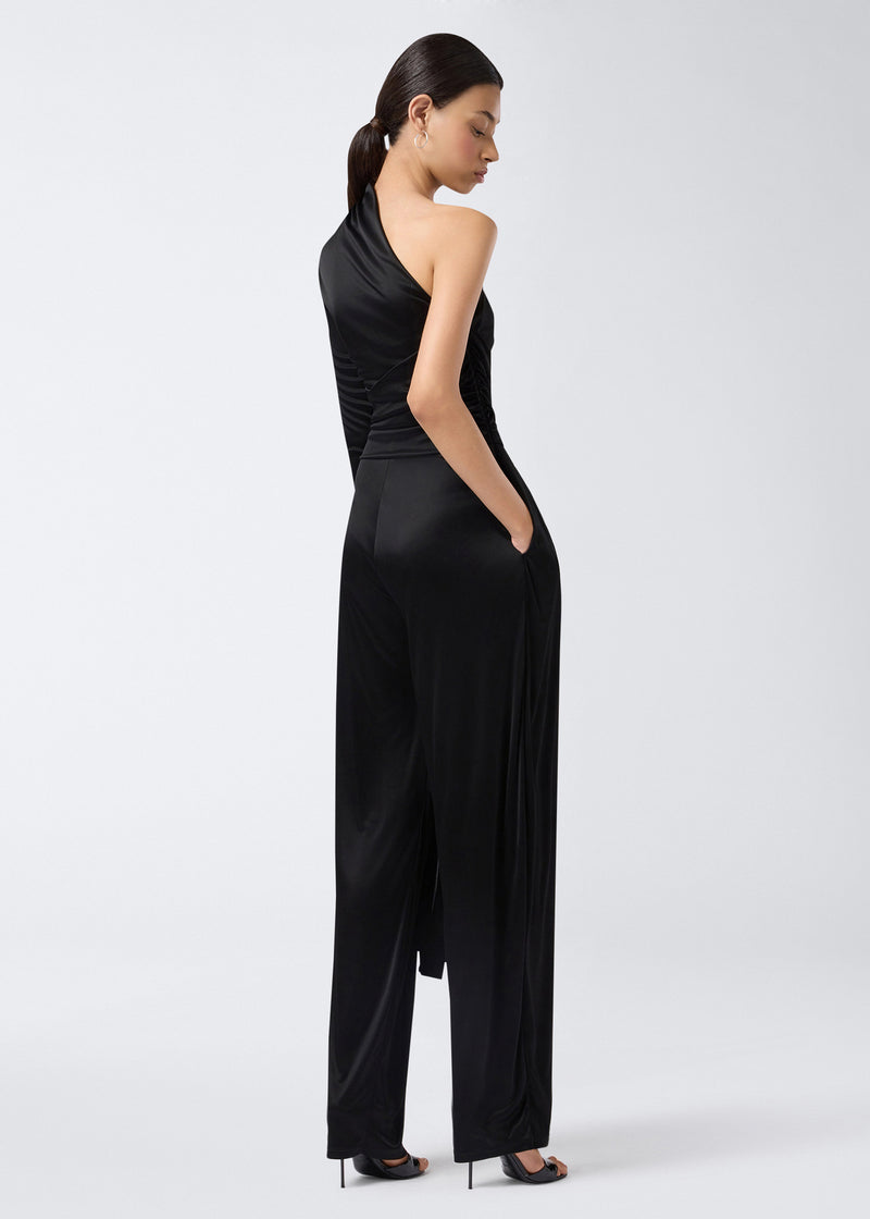 
                          Rear view of model wearing the Webster Jumpsuit, a black shiny jersey one-shoulder jumpsuit with a tonal waist belt and straight-leg pants.
                        