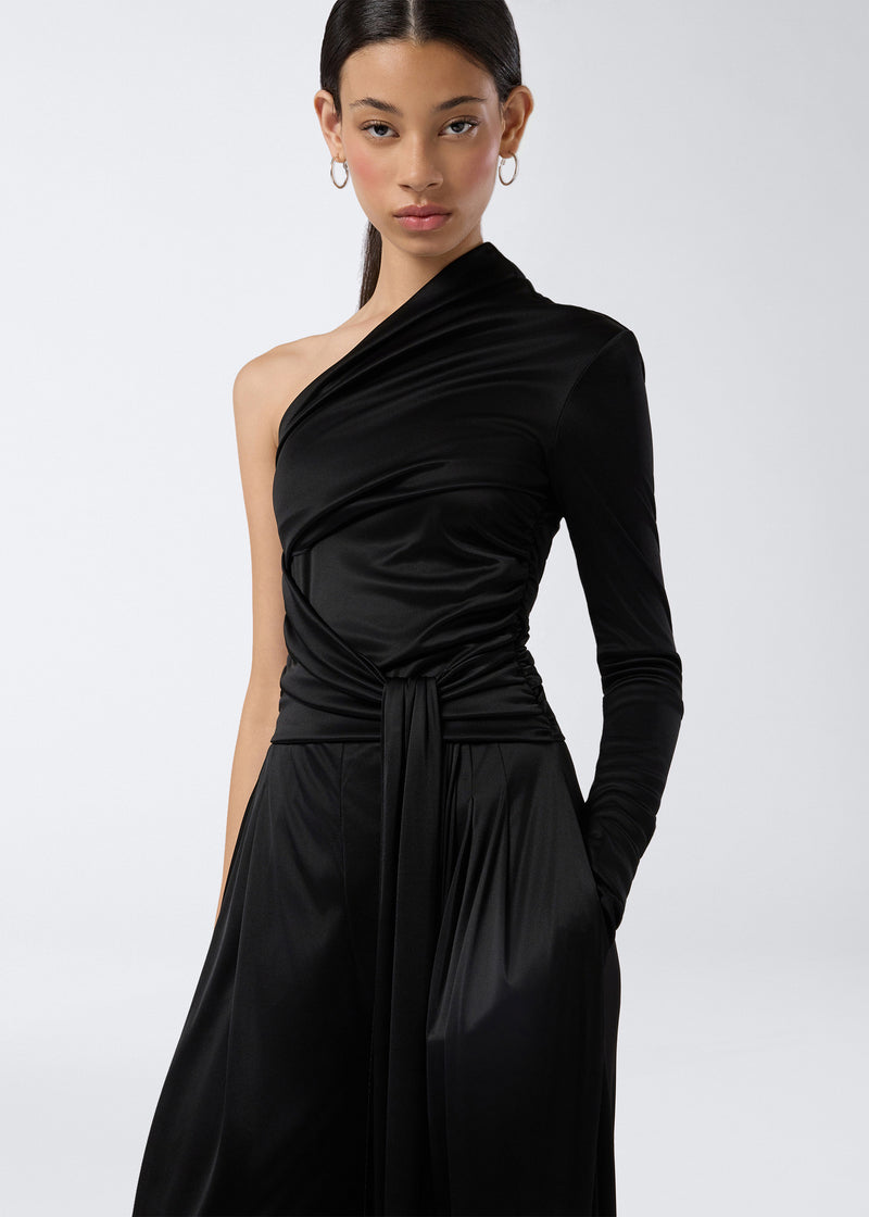 
                          Frontal view of a woman wearing the Webster Jumpsuit, featuring a black shiny jersey fabric, one-shoulder design, long sleeve, and a waist belt that drapes down.
                        