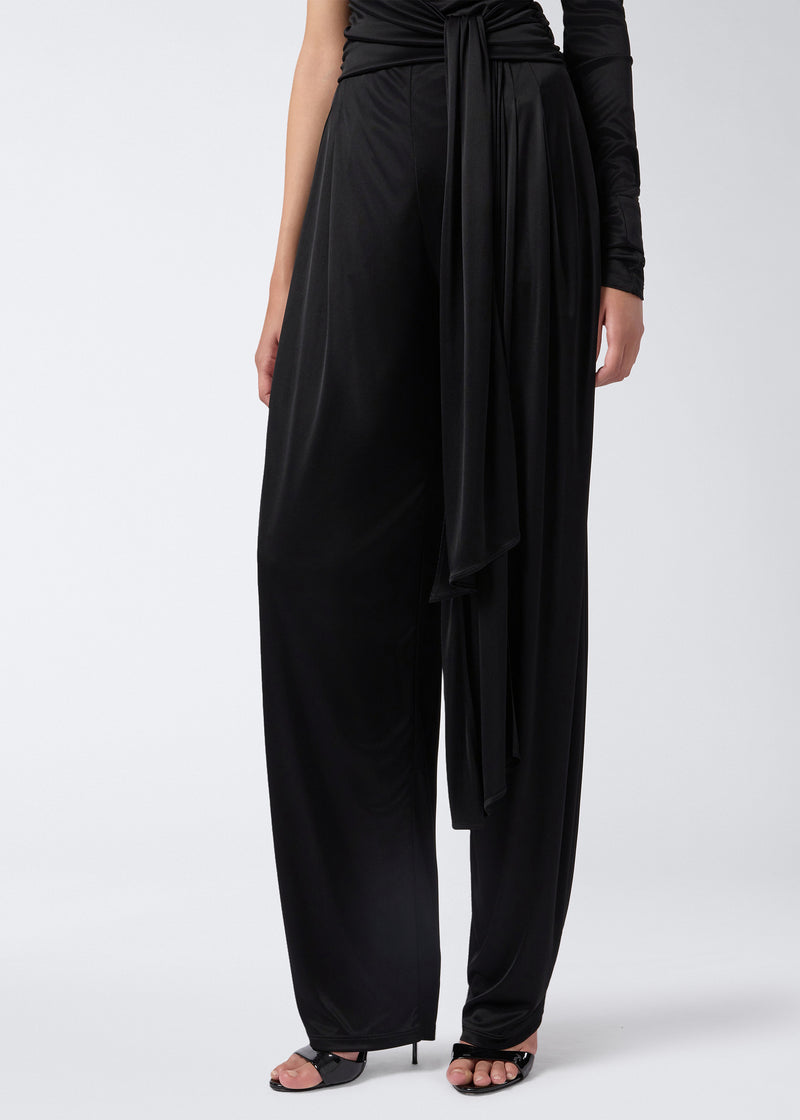 
                          Frontal view of the Webster Jumpsuit showing straight-leg black shiny jersey pants, a tonal waist belt draping past the knee, and one long sleeve.
                        