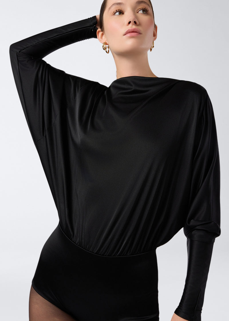 
                          Frontal view of a model wearing the Winnie Playsuit in black shiny jersey, featuring a boat neckline, long sleeves, and a fitted waist.
                        