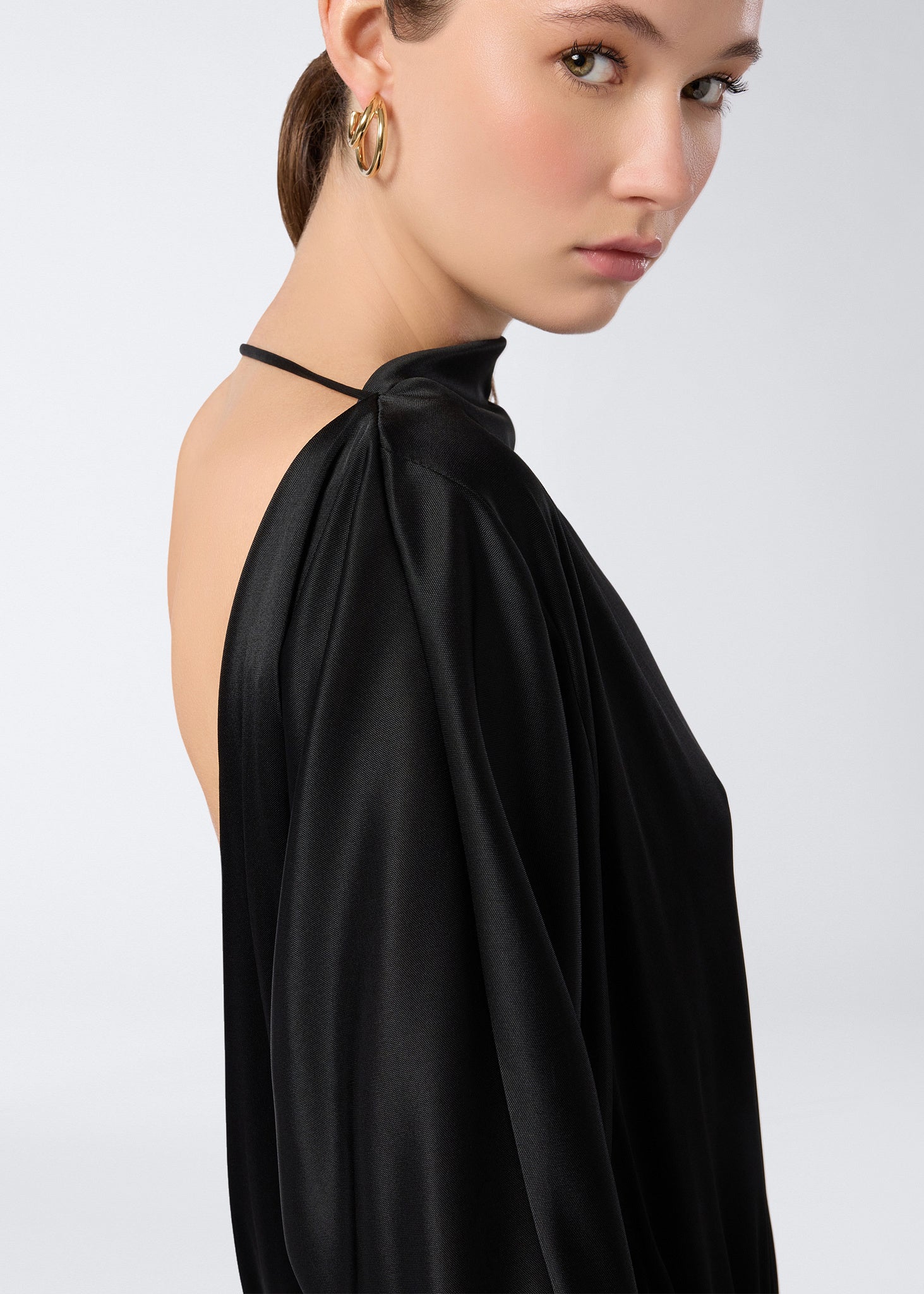Side view shows model wearing Winnie Playsuit in black shiny jersey with an open back, single neck band, and long sleeves, highlighting soft curved lines.