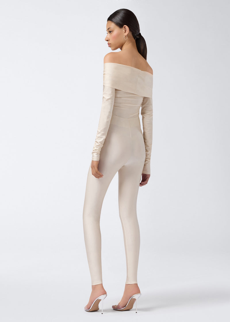 
                          Rear view shows a woman wearing the Kendall Jumpsuit in ivory shiny lycra, featuring off-the-shoulder long sleeves and fitted leggings extending to the ankles.
                        