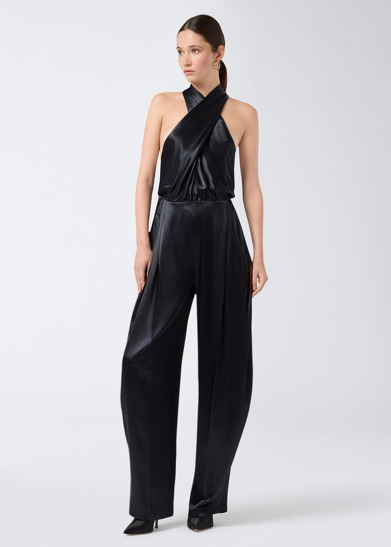 
                          Frontal view shows a model wearing the Zeyne Jumpsuit in black shiny satin, featuring cross-over halterneck straps and voluminous tapered balloon pants.
                        